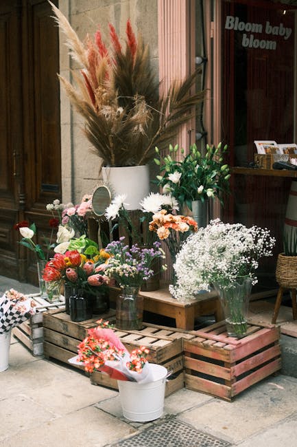 A variety of fresh flowers arranged in clear glass jars and vases outside a flower shop, featuring pink roses, white daisies, baby's breath, lavender, and peach-colored blossoms, complemented by tall