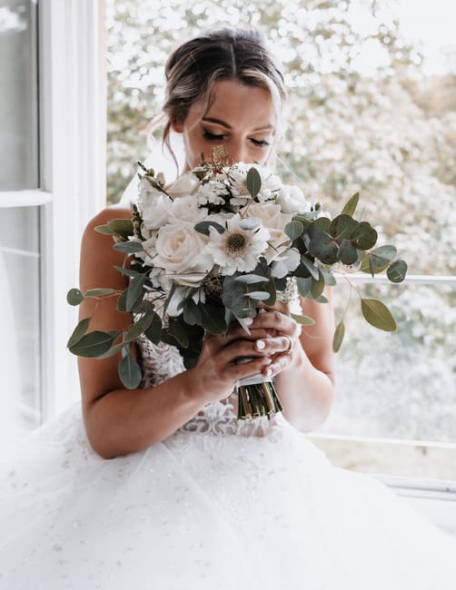 A bride holding a bridal bouquet featuring large white roses, cream-colored gerbera daisies, and sprigs of eucalyptus with round, silvery-green leaves. The bouquet is wrapped in a metallic gold ribbon