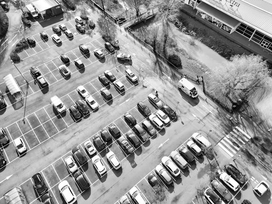 A black-and-white aerial view of a parking lot filled with numerous parked cars arranged in neat rows. The parking spaces are clearly marked with white lines, and some cars are casting shadows on the 