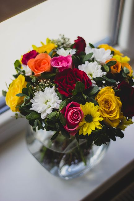 A vibrant floral bouquet arranged in a clear glass vase, featuring a variety of fresh flowers including bright yellow daisies, deep red roses, pink roses, white daisies, and yellow lilies, all accente