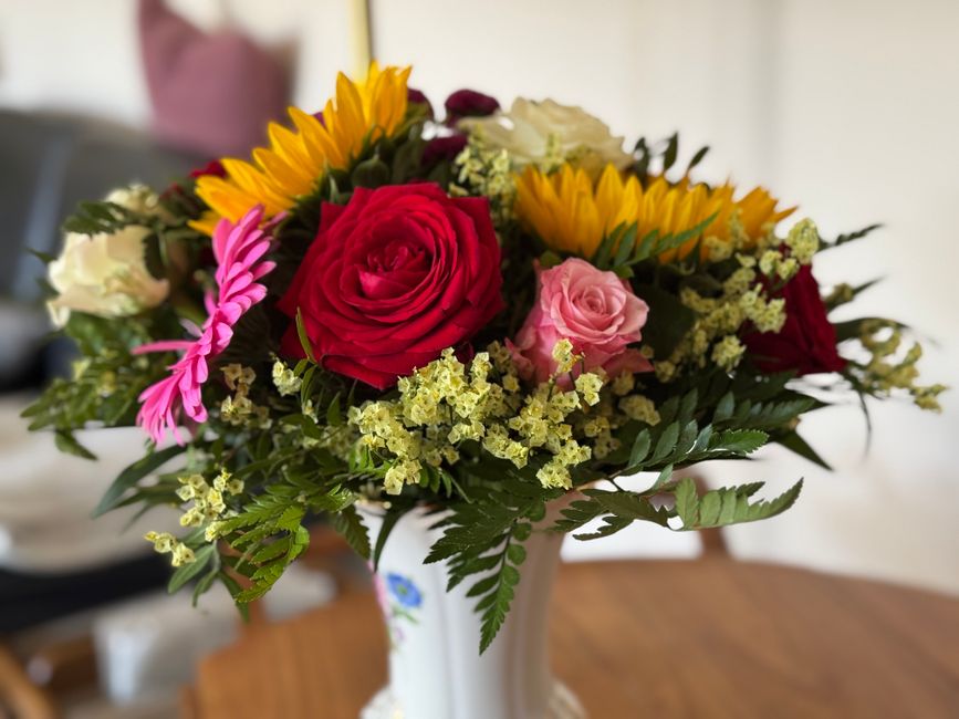 A vibrant floral bouquet arranged in a white ceramic vase, featuring red and pink roses, yellow sunflowers, pink chrysanthemums, delicate white baby's breath, and lush green foliage with fern-like lea