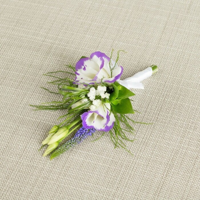 A refined and romantic boutonniere is photographed resting on a softly textured light beige linen surface, the gentle weave creating a calm, neutral backdrop that allows the colours and textures of the flowers to take centre stage. Dominating the composition are two beautifully formed lisianthus blooms, each petal a luminous white that curls outward in delicate ruffles, their edges dipped in a saturated purple hue that adds drama without overwhelming the eye. Tucked around these main flowers are small clusters of crisp white filler blossoms, reminiscent of baby's breath, which break up the strong bi-colour petals with a haze of softness and give the piece a light, breathy quality. Along one side runs a slender column of muscari, its tight, deep purple bells stacked like tiny beads, adding both vertical structure and a cooler, slightly blue-toned shade of violet to the palette. Two small, pale green buds extend from the arrangement, their closed forms hinting at fresh growth and the continuing life of the flowers beyond the wedding day. Behind and beneath the blooms, a mix of fine, feathery greenery and subtle leaves creates a soft cushion, the fronds suggesting movement and echoing the relaxed foliage you might see in the small gardens and hedgerows of North Watford. The lower portion of the stems is neatly gathered and wrapped in smooth white satin ribbon, the fabric catching light with a gentle sheen and tied off with a modest, tidy bow that signals careful craftsmanship. This close-up image conveys a sense of quiet sophistication and tenderness, suitable for a groom or groomsman heading to a ceremony in or around North Watford, and reflects the kind of thoughtfully balanced floral work local clients look for when planning weddings, proms, or formal celebrations.