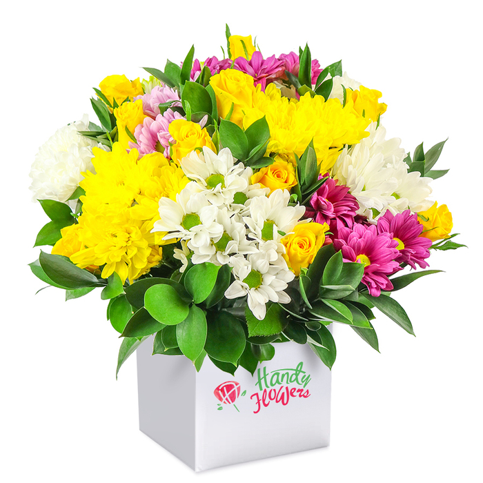 A densely packed, dome-shaped bouquet of fresh flowers sits in a sleek white square box, photographed against a spotless white backdrop so the colours look especially vivid. The arrangement is composed with the confident hand of an experienced florist from North Watford, balancing texture, height, and hue so that the bouquet appears both abundant and carefully controlled. Sunny yellow chrysanthemum heads, full and slightly ruffled, form much of the upper canopy, joined by clusters of small, tightly budded yellow spray roses that promise to open further over the coming days. Interspersed among them are clean white daisy-style chrysanthemums with pretty green centres, alongside larger, fluffier white mums that add a soft, billowing effect, like scattered clouds against a clear sky. Cutting through this harmony of yellow and white are vibrant fuchsia and magenta chrysanthemums; their rich tones, sometimes edged with lighter pink, offer a bold contrast that echoes the colourful seasonal displays you might spot in local North Watford gardens near the high street. Scattered rosebuds in gentle pink and sunny yellow nestle between the larger flowers, lending a sense of movement and life, as if the bouquet is still in the act of unfolding. At the base and throughout the arrangement, glossy dark green foliage provides a strong foundation, its sturdy leaves cradling the blooms and helping define the bouquet's rounded silhouette. The contemporary white cube case doubles as both container and gift packaging, making the arrangement feel ready to present for birthdays, thank-you messages, or to brighten a home or office. The even, bright lighting in the photo highlights the freshness of each petal and leaf, evoking the feeling of freshly prepared flowers waiting to be delivered across North Watford the same day.