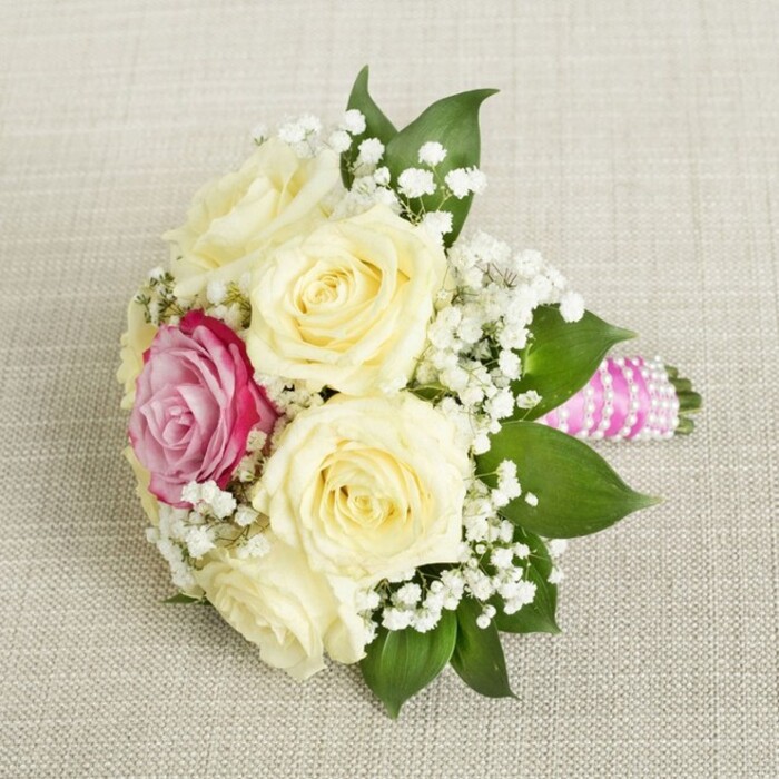 From an overhead angle, a compact floral bouquet commands attention, each detail meticulously arranged upon a neutral-toned, woven linen reminiscent of home interiors across North Watford. The main ensemble showcases five to six creamy, pale yellow roses, their petals still gently furled and bursting with fresh vitality-a visual embodiment of new chapters and quiet sophistication. Slightly off-centre, a single fuchsia-pink rose glows boldly, a vibrant contrast that instantly draws the gaze and connects the composition to the colourful flower stalls along Watford High Street. Sprays of delicate white baby's breath (gypsophila) float between the roses, lending a light, airy dimension that softens the display. Lush, glossy dark green leaves frame the outline, deepening the sense of natural abundance and providing a serene backdrop to the statement blooms. The handle emerges from the right, sheathed in a bright fuchsia-pink satin ribbon that shimmers in the gentle light, encircled by glowing white pearls wrapped meticulously in a spiralling pattern-every coil hinting at tradition and celebration. Resting atop a softly-textured, off-white canvas, the bouquet radiates understated elegance and joy, perfect for bridesmaids, intimate bridal moments, or refined North Watford events. The mood is one of thoughtful artistry and cheerful optimism, the design balancing modern vibrancy with classic romance-ideal for wedding planning inspiration, floral arrangement features, or discussions of sophisticated event florals in Hertfordshire.