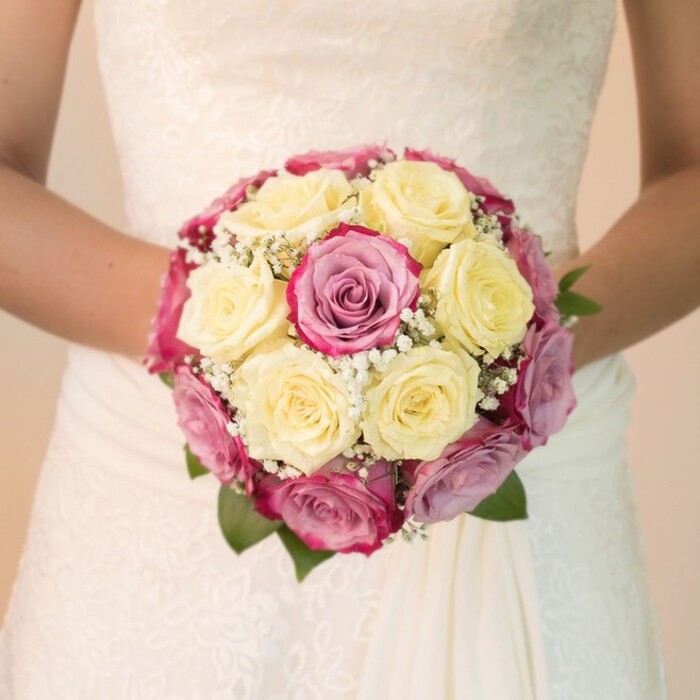 Set against the timeless elegance of a bridal gown, this meticulously arranged bouquet radiates the sophisticated charm found at weddings across North Watford. The focus lies on an exquisite assembly of roses: soft, creamy yellow blooms mingle seamlessly with vibrant fuchsia and hints of mauve rose petals, each flower skillfully positioned to create a balanced and inviting composition. Delicate sprigs of baby's breath thread through the arrangement like tiny lacework, their white blossoms catching the light with every movement. Subtle flashes of deep green leaves ensure the design remains grounded, while the bride's arms extend gently on either side, creating an intimate portrait of anticipation and joy. The gown's lace bodice, adorned with intricate floral motifs, reflects both tradition and contemporary grace, complemented by the gown's flowing skirt that drapes naturally beneath the arrangement. Natural daylight streams in, heightening the bouquet's freshness and lending each bloom and fold of lace a touchable texture. A softly blurred, neutral backdrop transports the viewer to a moment suspended in Watford's celebratory air-perhaps a sunlit ceremony near Oxhey Park or set in an elegant manor close to The Grove. In the hands of its wearer, this bouquet becomes a lasting symbol of commitment, crafted to celebrate love stories unique to North Watford.