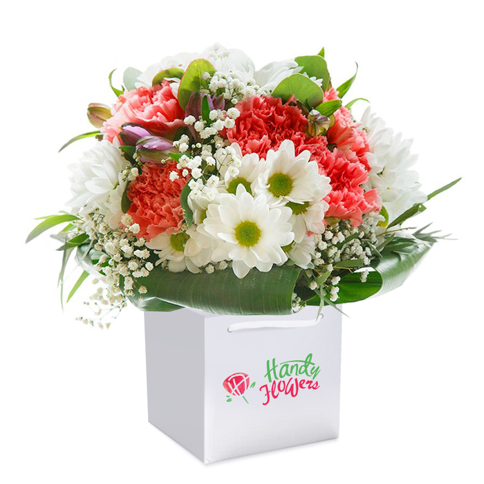 This image captures an exuberant floral bouquet standing upright in a sharp-edged white gift bag, perfectly staged against a pristine white background. The arrangement radiates freshness and skilled artistry; plush, coral-toned carnations with layers of delicate petals mingle with snowy white daisies that feature striking chartreuse centers-like rays of early daylight seen across North Watford's open greens. Scattered throughout are airy clusters of white baby's breath, their tiny blossoms filling the gaps and lending the whole bouquet a feather-like softness. Strategic pops of unopened buds, tinged with purple and edged in dusky pink, are evident on the left, providing both intrigue and a gentle promise of more blooms to come. Wrapping the base and sides, broad, glossy green leaves create a vivid contrast and a sturdy frame, reminiscent of the mature trees lining the residential lanes from St Albans Road to Leavesden Road. The bouquet itself forms a lush, full dome-thoughtfully balanced to capture both elegance and joyful abundance, whether chosen for birthdays, new beginnings, or spontaneous acts of kindness. The white gift bag, subtly stylish with white ribbon handles, suggests both sophistication and practicality, ready for hand-delivery anywhere in North Watford. Professionally photographed, each hue pops crisply against the uncluttered background, drawing the viewer's attention to the burst of texture and colour at the heart of the composition-showcasing the dedication of a North Watford florist to premium quality and community care.