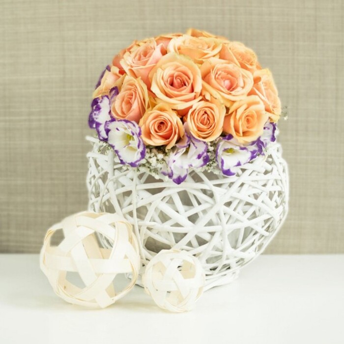 This image shows a carefully composed floral centerpiece that feels perfectly suited to an elegant wedding reception or anniversary dinner in North Watford. At the top of the arrangement, dozens of peach and apricot roses are packed tightly into a perfectly rounded dome, their petals overlapping in rich layers that suggest fullness and abundance. The roses appear at their peak, with softly curled edges that give them a plush, almost cushion-like texture. Just below, forming a gentle skirt around the rose dome, are clusters of white lisianthus blooms whose petals are finely outlined in a bold purple or violet hue. The coloured edges create a delicate frill that draws the eye around the entire circumference of the design. Threaded amongst them are tiny clusters of pure white gypsophila, their airy, cloud-like blossoms adding height and softness without disturbing the arrangement's balanced silhouette. The flowers are nestled into a striking white container made from thick, rattan-like strands woven into a bulbous, cage-like form. The open weave allows glimpses of shadow and negative space, giving the piece a sculptural quality. On the white surface beside the vase sit two additional decorative spheres, crafted from thin strips of pale, natural-toned wood, their open lattice construction echoing the main container's texture. Behind everything, a warm beige, gently textured fabric backdrop provides a calm, understated canvas that lets the subtle colours and craftsmanship shine. The mood is serene and sophisticated, ideal for a wedding breakfast at a North Watford venue, a styled photoshoot near North Watford station, or a refined dinner setting at home where guests can appreciate both the flowers' beauty and the artistry of the design.