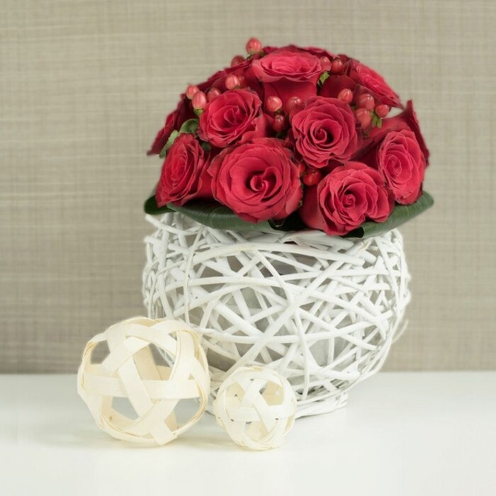 On a bright white tabletop, a striking red rose centerpiece draws the eye immediately, composed like a perfect wedding table arrangement ready for guests in North Watford. The flowers form a dense, rounded bouquet, with each rich crimson rose placed closely to its neighbour until the surface appears almost seamless. Some roses are at their fullest, revealing intricate layers of velvety petals, while others curl inward in half-open buds, creating subtle changes in texture and shadow. Scattered between the blooms are clusters of small red hypericum berries, slightly darker and glossier than the roses, giving the arrangement a nuanced, almost jewel-like detail. The base of the arrangement is discreetly wrapped in deep green foliage, just visible beneath the blooms, creating a soft collar that supports the weight of the bouquet. The flowers sit in a distinctive spherical vase made of white-painted twigs or wicker, woven into an open lattice that feels both handcrafted and contemporary. This airy structure lets the surrounding light filter through, casting delicate crisscrossed shadows on the smooth surface below. To the left of the vase, two decorative wicker spheres in a natural pale wood tone rest casually on the table, one larger and positioned in front, the other smaller and set behind, echoing the open-weave motif in a gentler, more rustic manner. Behind the scene, a softly blurred wall in warm beige or taupe adds quiet texture without distracting from the flowers. The image conveys a sense of refined romance and welcoming warmth, like a carefully styled wedding table at a North Watford hotel or local venue, ideal for celebrations, anniversaries, or romantic dinners at home.