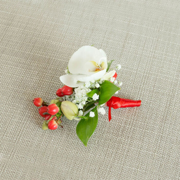 A single, carefully crafted boutonniere is displayed on a pale oatmeal-coloured fabric that looks like finely woven linen, the gentle texture forming an understated backdrop that allows every floral element to stand out with clarity. At the centre lies a striking white Phalaenopsis orchid, its petals wide, smooth, and gently overlapping, radiating from a warm heart of creamy yellow and soft orange tones that draw the eye inward. The orchid feels refined and contemporary, yet timeless, as if chosen by a couple marrying in one of North Watford's intimate venues or setting off from home for a registry office ceremony. Surrounding the central bloom, dainty sprays of white baby's breath create a soft, cloudlike fringe; each tiny blossom is crisp and bright, adding both volume and a sense of airiness that lightens the overall design. Just below the orchid, on one side, a compact cluster of glossy red berries adds a bold counterpoint, the rich scarlet tones gleaming like tiny ornaments, perfect for winter weddings, Christmas parties, or seasonal celebrations across the WD24 area. A few paler green buds sit nearby, still rounded and not fully open, hinting at ongoing freshness and life. Threaded between these details are small, verdant leaves in various shapes, some slightly pointed, others more oval, giving the boutonniere a natural flow and structure. At the base, all stems are neatly gathered and wrapped in a deep red satin ribbon, its sheen catching the light where it is tied into a snug, compact knot with short, tidy tails. The mood is polished yet heartfelt, a finely balanced combination of white purity, festive red, and natural greens that would suit a groom, groomsman, or prom guest setting out from North Watford with quiet confidence. Crafted with the same attention to detail that Flower Delivery North Watford brings to its local creations, the piece feels ready to be worn at a cherished occasion.