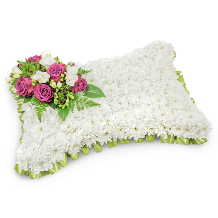 A formal, pillow-shaped sympathy arrangement set to soothe, fashioned from hundreds of tightly clustered white chrysanthemums that create a velvety, uniform surface across the rectangular form. The cushion-like mass, with softly rounded corners, reads like a peaceful textile of blooms - each chrysanthemum's round head contributing to a hush of white that suggests purity and quiet rest. Anchoring the composition in the upper-left corner is an elegant spray of deep magenta to fuchsia roses, their layered petals offering a warm, dignified contrast to the cool white field. Nestled amid those roses are sprigs of small white freesias or similar accent blooms, their delicate forms and gentle perfume lending tenderness and clarity. Beneath and around the accent cluster, assorted green foliage - including slender fern fronds and small glossy leaves - fans outward, providing depth, movement, and an organic frame that lifts the focal colors. The entire pillow is edged with a pleated light-green satin ribbon, the soft sheen of the ribbon peeking beneath the flowers to give a refined border that defines the silhouette without distraction. Set against a clean white backdrop and photographed under soft light that casts a subtle shadow, the tribute's three-dimensional quality is apparent and comforting. As someone who has prepared many such memorials for services across North Watford - from intimate chapel gatherings near St. John's Road to graveside services at local cemeteries - I recognise this piece as a traditional, respectful choice: a composed expression of love, remembrance, and the wish for peaceful rest.