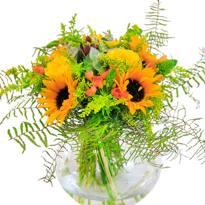 The first impression of this North Watford bouquet is pure sunlight captured in flowers-radiant sunflowers with bold, dark centres blaze alongside yellow roses, their petals opening in soft, intricate layers that glow like the early evening over Garston Park. Between these show-stoppers, sleek tendrils of greenery and cascading sprigs of fern tumble artfully, their varying textures and hues echoing the seasonal changes familiar to local gardens. Glossy red hypericum berries glint among the foliage, standing out like tiny gems against the matte softness of the petals. Eucalyptus leaves, tinged with blue-green, slip quietly between the brighter blooms, balancing the composition with cool, calming tones. Every vibrant stem is displayed through the clean, round glass vase, offering a glimpse of submerged green and the bouquet's graceful structure. Seen up close, one might imagine the subtle fragrances-roses with their gentle sweetness, eucalyptus's faint freshness, all blending into a scent as inviting as a North Watford summer breeze through Watford Market. This artful arrangement, available for same-day delivery across the area, brings an uplifting sense of joy-perfect for birthdays, anniversaries, or any local celebration where happiness is the guest of honour.