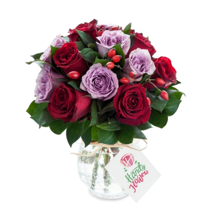 A carefully assembled, densely rounded bouquet of roses, where deep, velvety crimsons and somber burgundies meet a chorus of soft lavender and dusty purple blooms. Each rose is fully open, the petals unfurling in concentric layers that catch the light and reveal subtle tonal shifts - deeper edges, creamier inners - while the dark green foliage, likely Ruscus or Salal, provides a crisp architectural backdrop that highlights the roses' rich colours. Punctuating the arrangement are clusters of small, orange-red rose hips; their glossy, bead-like forms introduce rustic texture and a complementary pop of brightness that lifts the overall palette. The stems are gathered in a rounded mercury glass vase with an antique silvered, crackled surface that reflects and diffuses light, giving the display an elegantly aged shimmer; the vase narrows toward the top and is finished with a strand of natural raffia tied around the neck, from which a rectangular white tag hangs, stamped with a simple rose sketch and ready for a handwritten sentiment. Photographed against a clean white backdrop, the bouquet reads both luxurious and approachable, the kind of design a local florist might prepare for an intimate anniversary, a tasteful thank-you, or to brighten a bedside in a North Watford home. The visual impression is one of careful craftsmanship - tactile velvet petals, the firm give of berry skins, and the subtle, green-sweet scent of fresh foliage - a floral statement that balances romance and earthy charm.