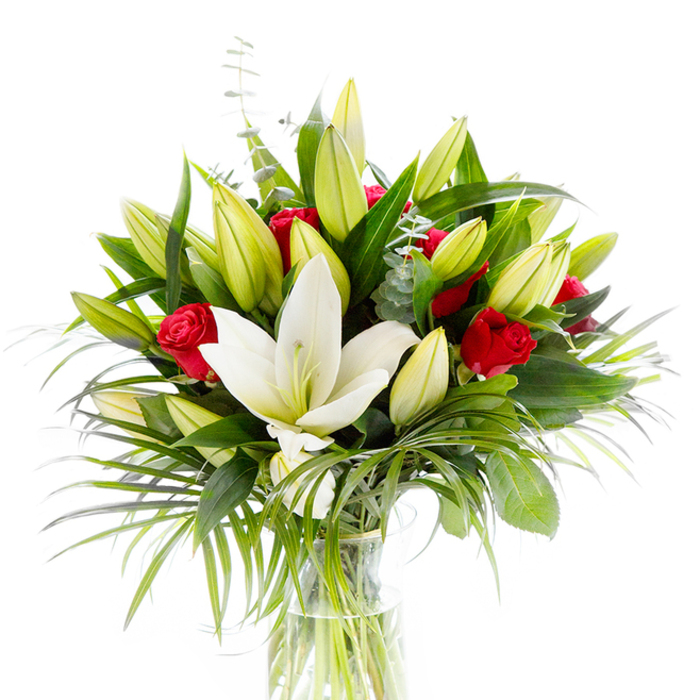 A striking, studio-lit bouquet arranged in a clean cylindrical glass vase and isolated on a white background, where every detail of form and colour is emphasised. At the heart sits one full, creamy-white Oriental lily, its satin-like petals curved back to expose pale yellow stamens and a delicate green pistil; around it cluster many lily buds in pale lime and soft yellow, suggesting unfolding promise and gentle motion. Interspersed among the lilies are several small, deep red rosebuds and partially opened roses, their velvety petals adding concentrated pools of passion and classic romance against the lighter tones. The green architecture is layered and deliberate: dark forest-green lily foliage gives the bouquet weight and silhouette, while lighter, feathery palm fronds sweep downwards and outwards to introduce a tropical, rhythmic flourish; silvery eucalyptus sprigs with rounded leaves soften the composition and introduce subtle tonal variety. The clear glass vase reveals tidy green stems in clear water, a small but honest sign of freshness and care. Under the bright backdrop the petals gleam with a soft sheen and the foliage casts gentle shadows, giving the piece depth without complexity. The overall impression is refined and celebratory - equally apt for anniversaries, Valentine's moments, or as a sophisticated centrepiece - and captures the kind of thoughtful design often chosen by customers in North Watford, from the Market area to residential streets near Cassiobury, who seek both purity and passion in a floral gift.