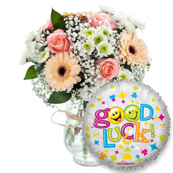 Set against a pristine white background, this cheerful good-luck arrangement balances gentle romance with playful celebration. A clear glass vase, finished simply with twine, cradles an abundant mix of pale pink roses and warm peach-orange Gerbera daisies whose dark brown centers lend a friendly contrast. Interspersed throughout are clouds of tiny white gypsophila and small crisp white-and-lime green chrysanthemum florets that add lively texture and a hint of fresh, green perfume; darker foliage threads through the blooms to create depth and movement. Light plays across the petals and glass, bringing out the soft, velvety rose tones and the Gerberas' satiny sheen. In the foreground, overlapping the lower right of the vase, a glossy silver Mylar balloon proclaims Good Luck! in a bubbly, multi-coloured font; the two o's are cheerful yellow smiley faces - one winking and one sticking its tongue out - while scattered hearts, stars and tiny geometric confetti in pink, blue, yellow and green animate the metallic background. The KALEIDOSCOPE logo is visible near the balloon's rim. Imagining a delivery route past Watford Market or a surprise left for someone strolling through Cassiobury Park, this composition reads as both intimate and celebratory - the kind of thoughtful, fragrant gift a local florist would send to encourage a friend starting a new job, taking exams, or embarking on any hopeful new beginning in North Watford.