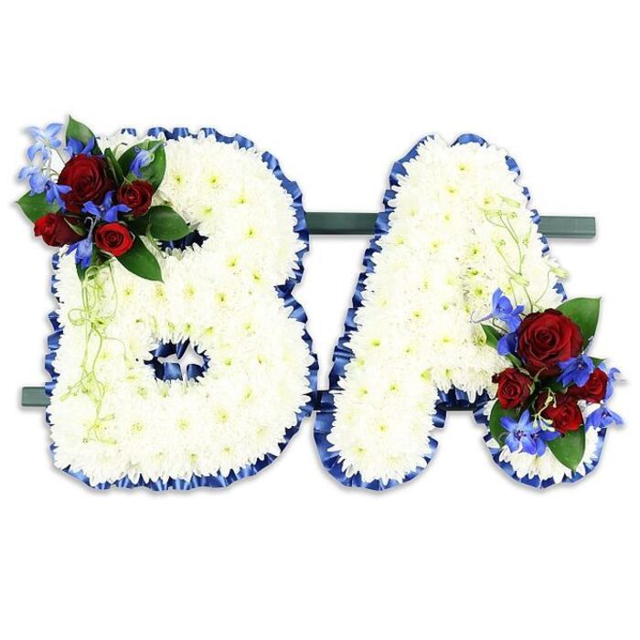 A carefully composed double-initial floral tribute, the capital letters 'B' and 'A' presented together on a narrow, dark green rectangular stand and photographed against a clean white backdrop. Each letter is meticulously covered with hundreds of tightly set white chrysanthemum florets, creating a uniform, cloud-like texture that conveys purity and quiet reflection. The edges of both letters are traced with a sumptuous royal blue satin ribbon, ruffled slightly to give definition and a formal, respectful frame to the delicate white surface. The 'B' bears an accent cluster on its upper left curve - a small, lush grouping of rich red roses in varying stages of bloom, their deep colour and soft scent providing a note of intimate remembrance. Sprays of vivid blue delphinium-like blooms are woven through the roses, their vertical, spiky shapes and electric hue adding contrast and depth, while darker green leaves and pale green trailing foliage tuck and spill gently around the cluster. The 'A' is similarly embellished at its lower right with a matching bouquet of red roses, blue accents, and mixed greenery, creating symmetry and a considered visual weight. Diffuse, natural light sculpts the chrysanthemums' soft surface and the ribbon's satiny sheen without harsh shadows, lending the piece a solemn dignity appropriate for a funeral or memorial service in a local North Watford church or chapel. The overall effect is traditional, personalised, and quietly expressive - a dignified floral message to honour a life.