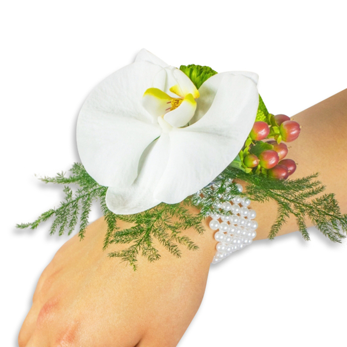 Set against a pristine white background, this wrist corsage rests on the light-skinned wrist of its wearer like a small, wearable garden crafted for a special day. The centerpiece is a large white Phalaenopsis orchid whose broad, satiny petals unfold with graceful simplicity; at its heart the labellum glows a vivid yellow with deeper orange-red accents, offering a concentrated warmth that brings the bloom to life. Beneath and around the orchid, delicate asparagus fern fronds unfurl in bright green, providing a soft, feathery bed that lifts the flower and introduces gentle movement. To the upper right of the bloom sits a compact cluster of Hypericum berries, transitioning in hue from pale green through peachy rose to a rich cranberry, their glossy skins and round forms adding weight and seasonal charm. A small section of ruffled lime-green foliage next to the berries introduces a fresh, contemporary highlight.

The arrangement is attached to the wrist by a refined multi-strand pearl bracelet that wraps softly and elegantly, the tiny white beads glimmering with an heirloom feel while remaining comfortable to wear. The lighting is calm and even-studio daylight that reveals texture without harsh shadow-so you can see the orchid's subtle veins, the fern's filigree, and the berries' polished sheen. As a local florist in North Watford, Flower Delivery North Watford often pairs classic blooms with modern accents like this corsage for weddings, Proms or intimate ceremonies, and the design reads as timeless yet fresh. Whether carried into a reception near Watford Junction or worn for portraits in Cassiobury Park, it projects understated luxury and thoughtful craftsmanship.