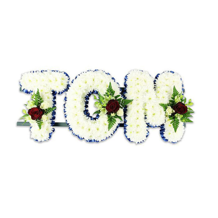 A striking, personalized memorial arrangement spells TOM in bold, soft letters made from densely packed white chrysanthemums; the small heads of the chrysanthemums create a continuous, textured face that reads both pure and substantial. Each letter's perimeter is sharply edged with a royal blue ribbon, the colour providing a refined border that visually separates the white floral mass from its surroundings and lends a formal, respectful tone. Inlaid into the center-lower areas of the T, O and M are symmetrical accent sprays: at the heart of each spray sits a single deep red rose, chosen for its rich hue and symbolic depth, surrounded by smaller ivory spray roses and tender green fern fronds that introduce contrast, rhythm and subtle movement. The letters are connected and stabilized by a thin, dark metal framework running just behind the base-this practical support keeps the tribute upright during viewing and procession. Set against a bright white backdrop and photographed in soft, neutral lighting, the arrangement's forms, textures and colours-white chrysanthemums, royal blue ribbon, deep red focal roses, ivory buds and fresh greenery-are clearly articulated. The overall effect is dignified, handcrafted and deliberately composed for a memorial setting. As someone who creates funeral tributes for families across North Watford, from Clarendon Road to the residential streets near Cassiobury Park, I would present this piece as a calm, elegant expression of sorrow and remembrance, suitable for chapel display or placement at a graveside.
