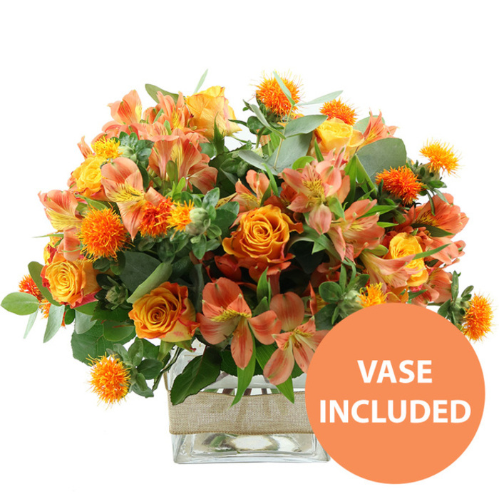 A generous, autumn-inspired bouquet in glowing oranges and yellows is captured in sharp detail against a spotless white background, as if it has just been set down in a bright North Watford living room. At the centre, several large orange roses form the focal point, each bloom perfectly opened to reveal concentric rings of petals that shift from deeper burnt orange at the outer edge to a softer, almost golden hue closer to the heart. Intermingled with these are sunny yellow garden roses, their densely layered petals giving them a plush, almost cushion-like look that adds both volume and elegance to the design. Slender stems of alstroemeria weave throughout the arrangement, their peach and soft orange petals gently curling back to expose fine speckles and subtle streaks of deeper colour, creating a painterly, almost watercolour effect within each tiny trumpet-shaped flower. Nestled between the larger blooms, small, spherical, spiky orange accents - reminiscent of safflower or thistle heads - punctuate the bouquet like tiny bursts of fireworks, adding texture and visual rhythm. A mixture of eucalyptus and other lush greenery surrounds and supports the flowers, the blend of silvery-green and darker foliage framing the warm tones so they stand out like autumn trees along the paths near Cassiobury Park. The stems are gathered cleanly in a clear, square glass vase, the water line visible and the stems arranged neatly for a sense of freshness and order. Around the centre of the vase, a broad band of natural beige burlap or jute is wrapped securely, lending a rustic, handcrafted touch that feels right at home in many North Watford houses and offices. In the bottom right corner of the image, an eye-catching circular orange icon bears the words VASE INCLUDED in bold white letters, making it instantly clear that this is a complete, ready-to-display floral gift, ideal for marking birthdays, anniversaries, or just brightening someone's day locally.