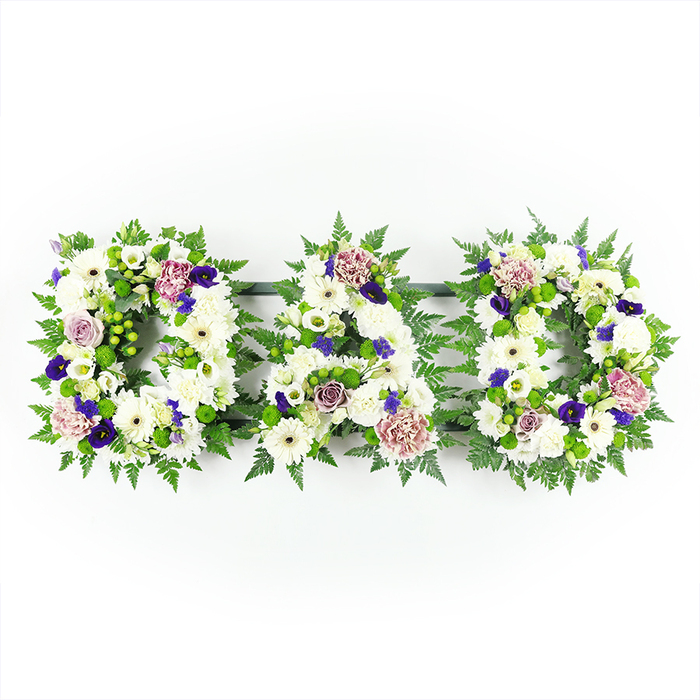 Three bold floral letters arranged in a gentle horizontal arc carefully spell out DAD, forming a heartfelt funeral tribute set against a pure white background that keeps all attention on the flowers themselves. Each letter is outlined and filled with abundant seasonal blooms, primarily snowy white chrysanthemums and daisy-like gerberas whose rounded petals and soft textures create a feeling of calm and purity. Tucked between them are smaller white roses and lisianthus, their layered petals opening modestly to suggest tenderness and remembrance. The structure of the letters is defined by generous greenery: fine, airy fern fronds and other lush foliage trace the contours of each D and the central A, giving the word a clear, recognisable shape and a vibrant green frame. Dotted throughout this white and green canvas are carefully placed accent flowers in shades of purple, lavender, and soft mauve. Deep violet blossoms reminiscent of pansies or small violas, as well as purple spray chrysanthemums or carnations, bring a sense of depth and dignity, while paler lavender roses and dusty pink or mauve carnations soften the look and introduce a gentle warmth. The composition is balanced and symmetrical, with each letter harmonising in colour and texture, like a single, unified statement of love. The lighting appears bright yet soft, with no harsh shadows, mimicking the clear daylight that might filter into a North Watford chapel or funeral home. This DAD tribute, the kind a skilled team at Flower Delivery North Watford would create for local services and burials, offers a deeply personal way to say goodbye, evoking quiet reflection and enduring affection for a beloved father.