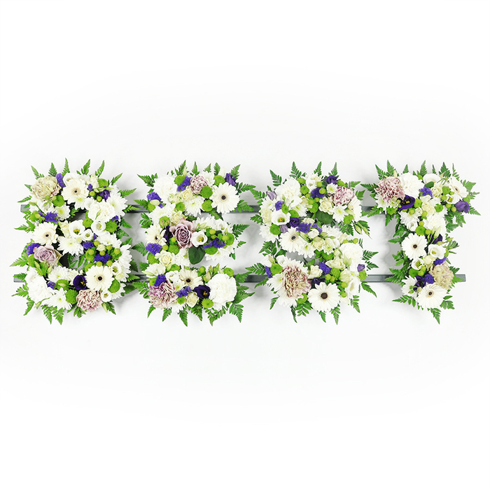 A broad, horizontal floral display spelled BEST, each uppercase letter shaped with meticulous care against a pristine white studio surface. The letters are woven from predominantly white flowers - large, immaculate gerbera daisies with their darker centres, delicate lisianthus, and layered white carnations that give a mix of smooth and billowy textures - evoking purity and gentle remembrance. Accenting these are soft lavender and mauve roses and carnations, plus smaller clusters of deeper violet filler blooms that add contrast and a warm, respectful undertone. The edges of each letter are lined with abundant green fern fronds, their feathery forms creating a soft cradle for the flowers and lending the tribute a sense of life and quiet movement. Scattered within are compact green button chrysanthemums that bring fresh, verdant highlights and three-dimensional depth, while the letters rest upon a dark grey to black structural base that peeks through in places to define the clean typography. The overall composition feels measured and tender, lit with soft, natural light that emphasizes petal textures and shadowed contours. This type of custom sympathy lettering is commonly chosen for memorial services or funeral tributes across North Watford, and at Flower Delivery North Watford we craft such pieces with careful selection of blooms and sensitivity to families' wishes, mindful of local venues from Watford General to small chapels near St Marys. The arrangement speaks of respect, lasting affection, and a composed celebration of a life.