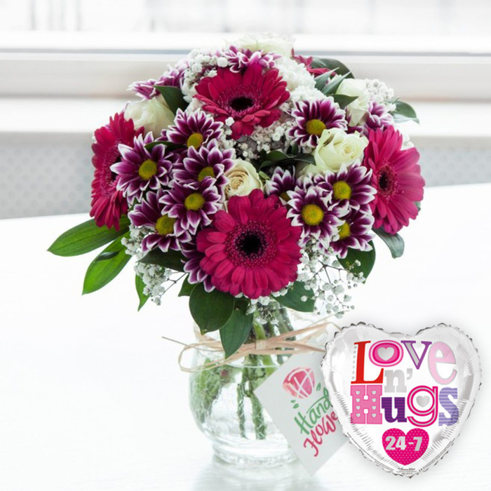 A lavish, celebratory bouquet takes centre stage in this image, as though it has just been placed on a dining table in a North Watford home after a same-day delivery. The arrangement bursts with colour, dominated by several large gerbera daisies in vivid fuchsia and magenta. Their wide, daisy-like faces are framed by layers of smooth petals and anchored by dark, velvety centres that add a dramatic focal point. Nestled around them is a generous scattering of smaller chrysanthemums, their petals painted in deep purple that fades to crisp white tips, forming a starburst around bright yellow centres that glow cheerfully against the magenta tones. Soft, creamy white roses are tucked among the bolder blooms, their gently curving petals giving the bouquet a refined, timeless elegance. Fine, cloud-like sprays of white baby's breath drift through the arrangement, filling spaces and bringing a delicate, airy texture that softens the overall look. Rich green leaves and foliage form a fresh, vibrant frame, supporting the flowers and rounding out the composition so it feels lush and complete. All of these stems are gathered into a short, clear glass vase, allowing a full view of the cleanly cut green stems and clear water, evidence of careful preparation by a professional florist familiar with local tastes. Around the vase neck, a simple band of natural-coloured twine is tied, from which hangs a small white tag adorned with a pink rose illustration and green script text that looks like it has been handwritten with care. To the right of the vase, a metallic heart-shaped balloon with a reflective silver edge leans cheerfully outward. Its white face is decorated with the phrase Love n' Hugs 24-7, the letters formed from playful patterns in shades of pink, purple, and blue, dotted with little hearts. The softly blurred, bright background suggests daylight streaming through a window, evoking the feeling of a thoughtful gift sent to share affection, mark an anniversary, or brighten a special day anywhere around North Watford.