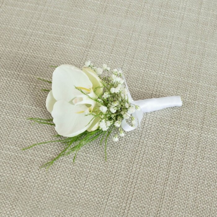 A meticulously crafted white orchid boutonniere lies elegantly on a pale beige, linen-like fabric, its woven texture visible beneath the flowers like a gentle backdrop often seen in North Watford homes and small wedding venues. The central bloom is a single, immaculate Phalaenopsis orchid, its petals thick, velvety, and perfectly formed, unfolding in smooth arcs of pure white. At the heart of the orchid, where the petals meet, a whisper of buttery yellow and subtle cream adds dimension, hinting at the natural complexity of a living bloom. Surrounding this focal flower, a generous collar of baby's breath erupts in a cloud of tiny white stars; each gypsophila head is delicate and round, creating a soft halo that contrasts beautifully with the orchid's bold, sculptural shape. Slender threads of vivid green foliage peek out from beneath the blossoms, their fine, feathery structure suggesting plants like asparagus fern or a similarly delicate greenery, as if freshly snipped from a garden near Garston or along the quieter streets branching off St Albans Road. These elements come together at the base, where the stems are neatly aligned and expertly wrapped in white satin ribbon. The ribbon is smooth and slightly lustrous, gathered snugly around the stems before being tied into a small, graceful bow that indicates the piece is ready to pin onto a suit jacket. Soft, even lighting falls across the boutonniere, casting minimal shadows and enhancing the gentle contrasts between the creamy white petals, the airy gypsophila, the vivid greens, and the warm neutral background. The overall mood is one of quiet sophistication and modern classic style, ideal for a groom wanting a minimalist yet luxurious touch as he prepares for a ceremony close to home in North Watford.