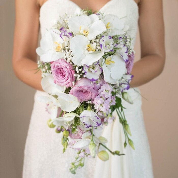 Set against a placid beige background evoking the tranquil streets of North Watford, this striking, vertical close-up captures a bride from the chest down, cradling an opulent, cascading bouquet. Crafted by expert local florists, the bouquet embodies a harmonious flow, with large, velvety white phalaenopsis orchids opening boldly amidst pastel pink garden roses-classic and full-petaled, radiating traditional romance. Soft clusters of lavender and lilac-toned stock bring gentle volume and whispering movement, as delicate white lisianthus with frilled petals-and the occasional purple-tinged edge-introduce a touch of contemporary flair. Wispy sprays of baby's breath glide throughout the bouquet, their cloudlike texture filling spaces with lightness, while threadlike sprigs of greenery add organic definition. The bouquet's teardrop shape gently cascades over the bride's simple, ivory lace strapless dress, whose intricate bodice and skirt detailing glimmer in the soft natural light. Tenderly held, the flowers seem to spill downward, echoing the gentle flow of North Watford's River Colne. This visual celebration invites the senses into a world of subtle scent, soft textures, and nuanced colour: blush, white, lavender, and gentle green. The craftsmanship shown in this arrangement highlights local wedding elegance, offering inspiration for those seeking a socially inspired yet timeless bridal accessory-perfect for North Watford couples wishing to keep their love story rooted in local tradition and floral expertise.