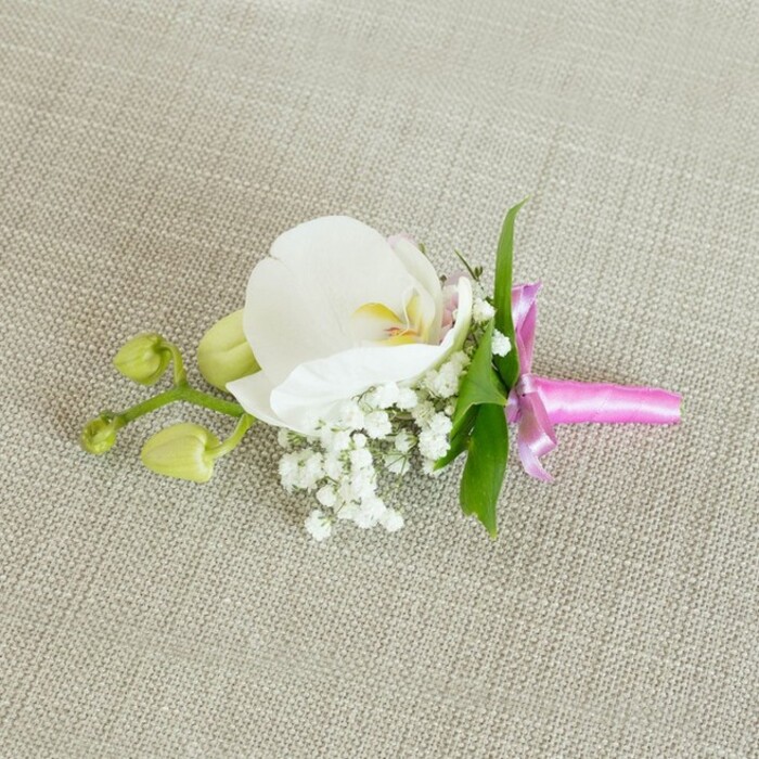A refined orchid boutonniere lies gracefully across a pale oat-coloured linen surface, the fabric's gentle texture lending an understated sophistication that would suit a North Watford ceremony or reception along St Albans Road. At its centre, a single white Phalaenopsis orchid unfurls, the petals broad, smooth, and almost porcelain-like, curving outward in soft, rounded arcs. The bloom's core reveals delicate notes of yellow and a blush of soft pink, drawing the eye inward to its intricate throat, where the fine markings suggest care and craftsmanship. Clustered at the base of the orchid, airy sprigs of white baby's breath create a cloud of tiny blossoms, their multitude of miniature blooms enhancing the sense of lightness and romance. Nestled among them, a few slim, vibrant green leaves stretch out, providing a fresh, natural contrast to the surrounding whites and echoing the greenery found in nearby gardens and parks. Off to one side, a slender offshoot carries a pair of closed, lime-green orchid buds, their smooth, rounded forms suggesting new chapters and quiet anticipation, as though the day's celebration is just beginning. The stems are carefully gathered and bound with a narrow satin ribbon in bright fuchsia pink, tied into an elegant little bow whose glossy sheen catches and reflects the soft, diffused light. The interplay of textures-the satin ribbon, the orchid's velvety petals, the tiny orbs of baby's breath, and the slightly coarse linen-creates a harmonious composition that feels both modern and timeless. Crafted with the precision of an experienced local florist, this floral accessory would sit beautifully on a groom's lapel, a prom suit, or a special guest's outfit, adding a discreet yet meaningful touch of colour and elegance to celebrations across North Watford.