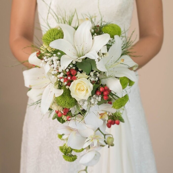 Captured in intimate detail, this photograph showcases a bride in North Watford holding a lush, cascading bridal bouquet against the subtle ivory lace of her wedding gown. The central focus is a striking interplay of large, trumpet-shaped white lilies-each marked with gentle, russet freckles-surrounding a single, beautifully tight cream rose that echoes classic romance. Delicate white Phalaenopsis orchids spill down from the ensemble, their slender stems creating an elegant, elongated teardrop shape that enhances the bouquet's movement. Airy clouds of baby's breath interlace among the larger blooms, softening the ensemble and lending visual lightness, while clusters of vivid lime-green pom mums and glossy red Hypericum berries provide energetic bursts of colour and texture. Long, wispy foliage frames and supports the composition, lending an organic touch that evokes the gardens and parks of North Watford. The bride's hands, delicately holding the ribbon-bound stems, disappear into the gentle blur of her softly lit dress, allowing the florals to take center stage. This sophisticated arrangement, a favourite among local North Watford couples, exemplifies the region's affinity for both traditional elegance and modern flair, making it perfect for articles about contemporary bridal styles and wedding flower artistry.
