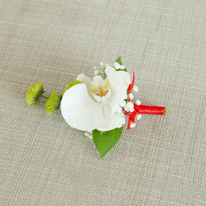 A single, elegant boutonniere lies thoughtfully arranged on a softly woven, light beige fabric that resembles natural linen, its understated texture and neutral tone echoing the gentle charm of a North Watford registry office celebration. At the heart of this refined floral piece sits a large white phalaenopsis orchid, its petals broad, velvety, and perfectly formed, opening like calm, symmetrical wings. The orchid's centre offers a delicate burst of colour: buttery yellow shading into fine strokes of reddish-orange and soft brown, creating an intricate pattern that draws you closer, even in a still photograph. Surrounding the central bloom, loose clusters of dainty white baby's breath are nestled in, their cloud of miniature flowers adding airiness and softness, like a light veil around the main blossom. On the left side, two rounded lime-green chrysanthemum buds stand out with their densely packed, spiky petals, adding an energising contrast in both texture and colour that feels contemporary yet still romantic. A generous, deep green leaf rests beneath the orchid, acting as a natural backdrop and giving weight to the design, while a smaller, darker leaf near the base provides subtle balance. The stems of the flowers are gathered neatly and bound with a vivid crimson red satin ribbon, wrapped tightly and finished with a tidy knot or small bow, introducing a vibrant ribbon of colour that suggests celebration and joy. The light falls evenly across the arrangement, highlighting the orchid's gentle sheen and the crisp weave of the fabric. Crafted with the level of care you would expect from a local North Watford florist, this boutonniere feels ideal for a groom, groomsman, or prom guest wanting a classic yet slightly modern floral accent, easily prepared for same-day local collection or delivery.
