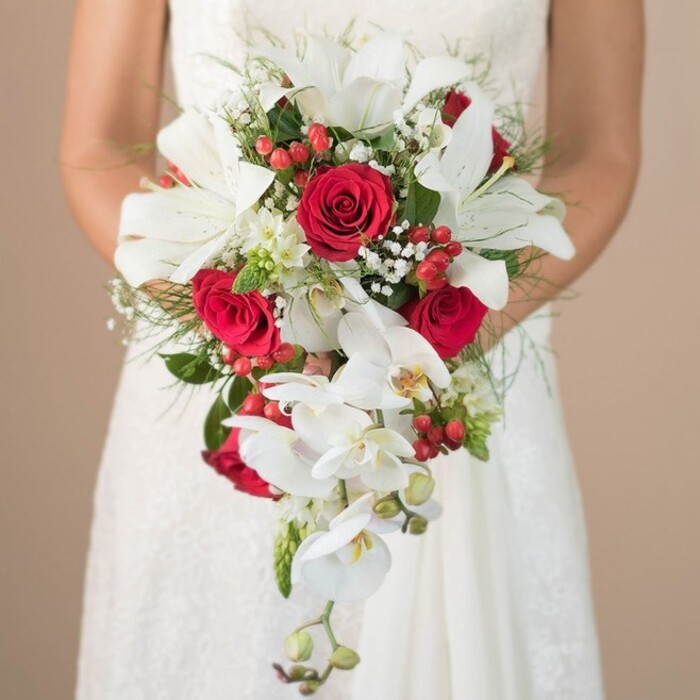 Capturing a moment of pure romance in North Watford, this close-up image frames a bride in an immaculate white gown as she gently holds a sweeping, cascading bouquet-designed to be the crowning glory of her wedding ensemble. The artistry of the bouquet is revealed in its harmonious blend of colour and form: bold, fragrant white lilies emerge as focal points, their broad petals radiant and luminous, contrasting beautifully with an array of opulent red roses nestled among them. The velvety richness of the roses hints at deep passion and enduring love, a fitting symbol for vows exchanged at St. Andrew's Church or a local ceremony. White phalaenopsis orchids create a delicate cascade, their petals and faint yellow centers whispering elegance as they trail gracefully downward, evoking the gentle rhythm of North Watford's leafy avenues. Peppered among these blooms are tufts of ethereal gypsophila, their miniature white flowers adding both volume and a dreamy, cloudlike softness. Bright red hypericum berries, glistening with freshness, pop alongside the blooms, adding playful visual interest and texture reminiscent of Watford's vibrant community gardens. Fine asparagus fern and polished deep green leaves complete the composition, giving structure and splashes of green reminiscent of local nature reserves. The bride's tender embrace of the bouquet, set against her detailed lace bodice, is bathed in soft, neutral light, focusing attention on the intricate arrangement. This bouquet, crafted by an expert North Watford florist, embodies both tradition and freshness, promising a memorable accent to any local wedding.