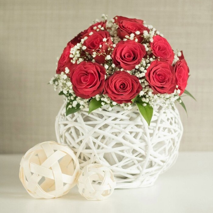 The image presents a romantic floral centrepiece designed with the precision and warmth of an experienced North Watford florist, ideal for dressing a wedding table or intimate celebration. A generous cluster of red roses forms a perfect, rounded mound, each rose open just enough to display layers of velvety petals that spiral inward in tight, luxurious swirls. The colour is a deep, classic crimson, evoking timeless love and quiet passion, like a bouquet prepared for a couple celebrating at a local venue near Cassiobury Park. Weaving softly among the roses are countless tiny blooms of white gypsophila, their fine stems and delicate flower heads adding a cloud-like lightness and an almost lace-like texture that plays beautifully against the richer tones. Small glints of green foliage peek between the roses, providing subtle contrast and suggesting freshness, as if the arrangement has only just arrived from a nearby flower shop. The bouquet is hosted in a distinctive spherical vase crafted from white, interlacing strands, forming an open, basket-like pattern that curves gently around the flowers. This woven design casts graceful shadows and creates airy gaps, giving the piece a handcrafted, artisanal feel. Resting beside the vase on a sleek, white tabletop are two decorative orbs made from pale, ribbon-like strips that twist into open spheres, one slightly larger than the other. These orbs echo the vase's lattice structure, tying the décor together in a cohesive, contemporary way. Behind everything lies a neutral, softly textured background in tones of warm cream or beige, intentionally understated so the eye is drawn to the interplay of red, white, and green. The arrangement feels both sophisticated and welcoming, perfect for evoking romance in a North Watford home or at a special event.