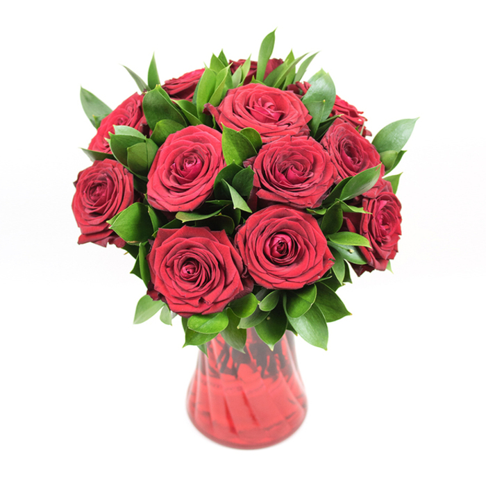 A richly romantic bouquet of approximately a dozen to fifteen deep red roses is displayed in a translucent red glass vase, all set before a crisp white studio background that heightens the drama of the colours. Each rose is at its peak, the blooms fully open but still brimming with life, their velvety petals arranged in tight spirals that gradually loosen toward the outer edges. The red tones are intense and uniform, evoking the classic language of love-something between garnet and crimson, with subtle variations where light brushes across the petal edges. The florist has shaped the arrangement into a perfectly rounded silhouette, every flower head carefully positioned so that the bouquet looks full from every angle.

Between the roses, clusters of glossy, dark green foliage-likely ruscus or a similar leaf-emerge and recede, adding layers and texture. The leaves are smooth, slightly pointed, and a rich, even green, creating a cooling contrast that makes the red appear even deeper. This greenery fills in the spaces between blooms, ensuring no gaps pierce the lush dome, giving the bouquet a polished, professional finish. The stems gather neatly into a red glass vase, whose gently flared neck and softly curving body echo the shape of the bouquet above. Its subtle ribbed or swirled texture captures fine highlights, while the transparency of the glass allows a suggestion of stems and shifting crimson tones within.

The white backdrop and even lighting give the scene a timeless, almost gallery-like feel-easy to imagine this same arrangement gracing a dining table in a North Watford family home near Garston or acting as the centrepiece in a modern flat overlooking the local parks. It speaks clearly of anniversaries, Valentine's Day, and quiet, devoted love: a simple, decisive gesture where red roses say everything that needs to be said.
