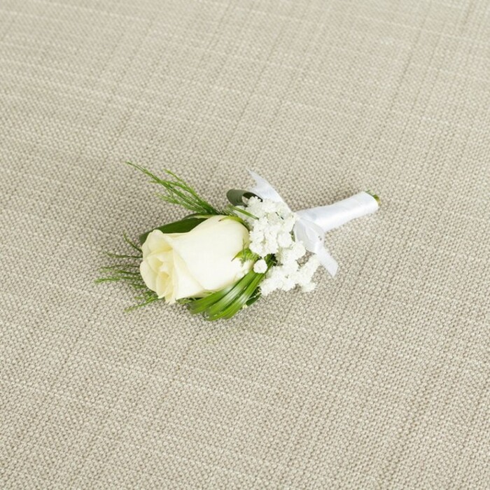 A single ivory-white rose boutonniere is captured in a still, contemplative moment, resting diagonally on a pale beige fabric surface whose finely woven, linen-like texture adds a natural, organic backdrop, much like the understated interiors of small North Watford venues along St Albans Road. The central rosebud is tightly formed yet softly rounded, with its outer petals just beginning to separate, giving an impression of new beginnings and careful timing, as though it has been cut at exactly the right moment by an experienced florist. Nestled close to the rose are airy clusters of white baby's breath, each spray composed of countless tiny blossoms that create a misty, cloud-like ring around the main flower, softening the silhouette and adding intricate detail for the fingertips as much as for the eyes. Sleek, deep green leaves and delicate, fern-like sprigs frame the arrangement, curving gently around the rose and gypsophila, their varying textures and tones reminiscent of the layered greenery in North Watford's private gardens and small public green spaces. All the stems are neatly gathered and wrapped in a band of smooth white satin ribbon, the wrap even and snug, culminating in a compact bow tied just under the flower head, its short loops and tails lying flat as though ready to be pinned to a groom's lapel for a ceremony at a local registry office or church. Soft, diffused lighting bathes the scene, casting minimal shadows while emphasizing the satin sheen of the ribbon, the velvety surface of the petals, and the grain of the background fabric. The boutonniere conveys an atmosphere of calm preparation and quiet romance, evoking that moment when someone in North Watford fastens it carefully in a mirror before stepping out to meet their future, carrying this small emblem of love and tradition on their chest.