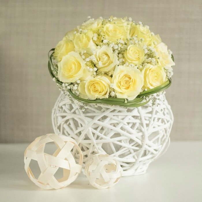 A graceful wedding-style table arrangement is captured in the frame, built around a generous cluster of pale yellow roses that form a rounded dome of velvety petals. Each rose seems to rest closely against its neighbour, creating a continuous surface of soft, buttery colour that glows gently, as if catching diffused daylight through a nearby North Watford window. Threaded delicately between the roses, white baby's breath unfurls in airy tufts, their countless tiny blossoms decorating the arrangement like fine lace and breaking up the mass of yellow with crisp white accents. Around the base of this floral mound, a narrow band of glossy green foliage has been neatly wrapped, adding definition and hinting at the florist's careful hand. Supporting the blooms is a distinctive white woven vase, curved subtly at the sides and crafted from interlaced strands that form an open, airy structure. Its pattern resembles modern wicker work, with narrow gaps that let the background hue peep through and cast gentle, linear shadows on the smooth white surface it rests upon. Nearby, two natural rattan-style decorative balls in a light wood tone echo the vase's weaving, their rounded shapes and open design tying the whole composition together. The neutral beige background is softly unfocused, like a calm wall in a North Watford home near the Dome Roundabout or along St Albans Road, ensuring the viewer's attention remains on the floral artistry. This arrangement evokes freshness, understated celebration, and the kind of timeless elegance suitable for wedding breakfasts, anniversary dinners, or refined home decor across the local area.