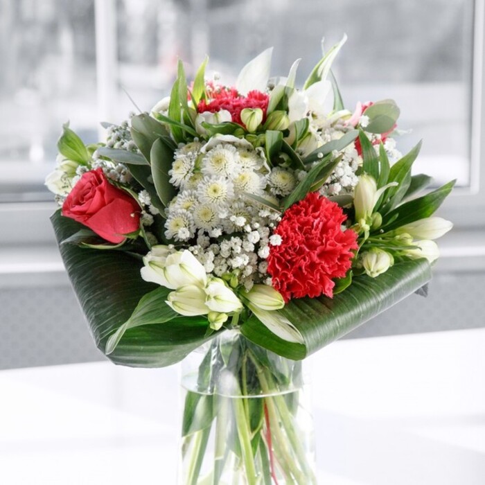 In this image, a sophisticated red and white floral arrangement rises from a clear cylindrical glass vase, the stems aligned in clean vertical lines beneath the water, hinting at the care and precision behind the design. Above, the main story is told in rich shades of red and crisp white. Deep red roses sit prominently, their velvety petals layered tightly at the centre and gently curling outward, evoking timeless romance and deep affection. At the front, a large crimson carnation acts as a focal point, its deeply ruffled petals creating ripples of texture that contrast beautifully with the smoother rose petals. Threaded between the bold reds are several types of white blooms: creamy white alstroemeria with elongated, slightly flared petals marked by fine speckles, soft daisy-like flowers that lighten the mood, and several upright white lily buds, still closed, suggesting the bouquet will continue to evolve over the coming days. Delicate sprays of gypsophila, or baby's breath, appear throughout like tiny snowflakes, lending a light, ethereal quality that softens the strong colour palette. The greens play an equally important role. Broad, glossy dark green leaves are wrapped and curled at the base, forming a sculpted collar that cradles the arrangement and conceals the stems just above the vase rim. Taller, more slender foliage, including eucalyptus-like stems and pointed leafy spikes, adds structure and height, suggesting the gentle movement of the trees lining the streets of North Watford. The bouquet rests on a smooth, white surface with a softly blurred, pale background that might be a sunlit wall or window, bathing the flowers in even, natural light. The overall atmosphere is calm yet celebratory, perfect as a refined centrepiece for a dinner in a Watford home, a thoughtful Mother's Day surprise, or a graceful congratulations bouquet delivered locally to mark milestones both big and small.