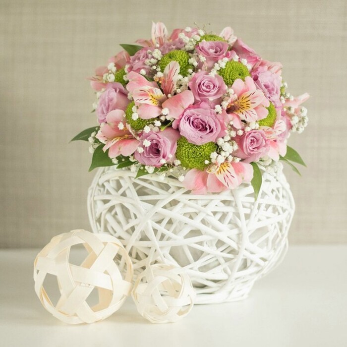 A radiant display of North Watford floral artistry is captured in this image: a centerpiece merging soft, romantic pastels and crisp greenery to achieve a statement of modern elegance. At its core, full-bloomed roses in shades of pastel pink and delicate lavender layer upon one another, their velvety petals shimmering with freshness. Interspersed among these are airy clusters of pink alstroemeria, known for their slender petals adorned with subtle streaks, adding gentle movement and variety to the round bouquet. Bright lime green button chrysanthemums, their pom-pom shapes playful and eye-catching, bring a spark of energy, while frothy white sprigs of baby's breath impart a dreamy, ethereal effect. The floral arrangement is nestled into a unique white vase: its surface intricately woven into an open, basket-like design, reflecting the creative, handmade spirit of florists serving North Watford's welcoming communities. The rounded form of the vase supplies visual and literal stability to the generous bouquet. Completing the composition, two decorative orbs-crafted from light, natural-toned reeds or rattan-are set to the left of the base, their varied sizes and open patterns harmonising with the vase's silhouette. The arrangement is lit with soft, ample lighting that highlights nuances in color and texture against a gentle, fabric-textured beige wall. This centerpiece is perfectly suited to adorn wedding tables in venues around North Watford or to bring an air of sophistication and heartfelt joy to local celebrations, capturing the essence of thoughtful design and of fresh, locally sourced blooms.
