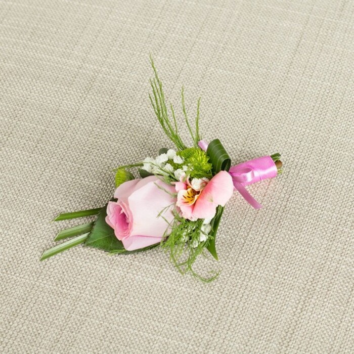 Nestled atop a subtly woven, ivory-toned linen, this wedding boutonniere captures the timeless romance of North Watford with every detail. Its centerpiece-a single, tightly furled rosebud in gentle pastel pink-projects an aura of delicacy and hope. Around the rose, textured elements are masterfully interlaced: the ethereal clouds of baby's breath, sprightly green mini-chrysanthemums, and a second, ruffled flower in blush-pink or peach, perhaps an Alstroemeria, with golden-yellow highlights and intricate central speckles. Rising from this arrangement, slender sprays of feathery grass and fern-like greenery arch gracefully, reminiscent of the natural foliage found in local parks. Lush, darker green leaves form a compact, folded base beneath the blooms, lending depth and structure without overshadowing the blossoms' soft hues. The stems are expertly bound with a vivid fuchsia satin ribbon, tied into a small, tailored bow-its vibrant colour bringing a celebratory note that contrasts beautifully with the boutonniere's gentle palette. The overall look suggests a contemporary classic perfect for grooms or prom guests across North Watford, emanating quiet elegance and a whisper of fresh garden scent. This floral pin would feel at home at a wedding venue near St Albans Road or at an intimate celebration at a family-run event space, embodying both local heritage and modern style.