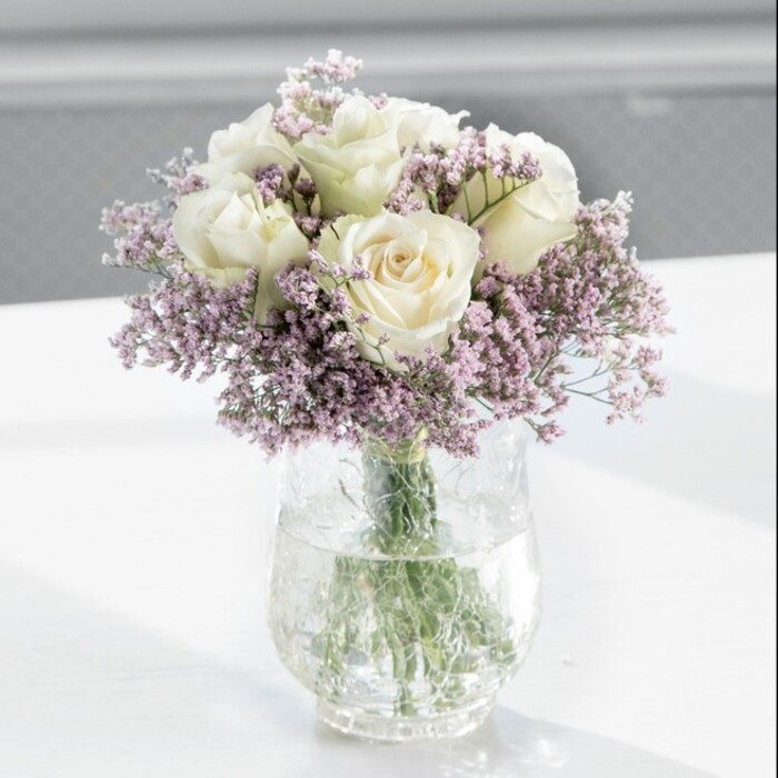 The image shows a classically elegant bouquet composed mainly of creamy white roses, accented with clouds of pale lavender statice, arranged with professional precision in a tall, clear glass cylinder vase. At the centre of the display, roughly a dozen white roses create a lush, domed shape, each flower at an ideal stage of openness: not tight buds, but not fully blown either. Their petals are velvety and gently curved, layered in soft spirals that draw the eye toward the middle of each bloom. The colour feels like warm ivory rather than stark white, lending a gentle glow that carries a sense of timeless romance and calm. Intertwined with the roses, fine stems of lavender-hued statice break up the cream with a veil of tiny, clustered blooms. These filler flowers, with their delicate, papery texture, create a light, airy halo around and between the roses, adding depth and dimension without overwhelming the design. All the stems are visible through the water inside the clear vase, showing tidy, freshly cut ends and crisp green tones that hint at a bouquet prepared the same day, perhaps at Flower Delivery North Watford before being sent to a home near Garston or a quiet office along St Albans Road. The vase rests on a smooth, white surface that reflects some of the soft daylight washing into the scene. The background is gently out of focus, suggesting a bright, modern interior with large windows somewhere in North Watford, so that the light seems to fall naturally across the flowers, lifting every detail. The arrangement feels appropriate for many occasions-sympathy, anniversary, or a refined housewarming gift-bringing a mood of peaceful grace and dignified affection.