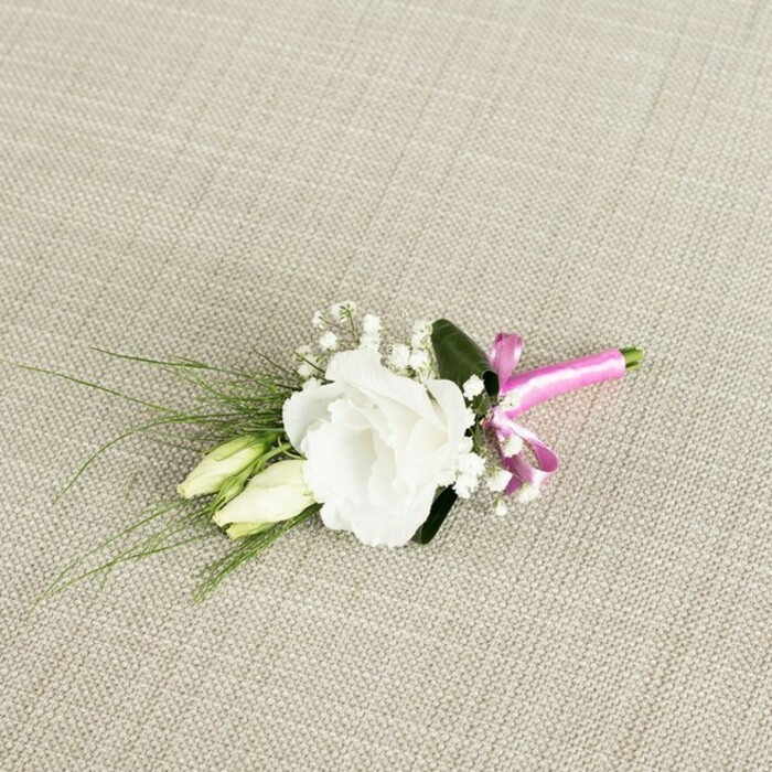 Set against a softly textured backdrop of pale beige fabric, reminiscent of natural linen spread over a wooden table in a North Watford townhouse, a single, handcrafted boutonnière is captured with gentle, even lighting that highlights every delicate detail. At the centre lies a fully opened white lisianthus flower, its many ruffled petals unfolding in graceful layers, creating a rounded bloom that looks almost like a miniature garden rose. The petals are a pure, clean white, subtly shaded by the light to reveal their velvety texture and fine edges. Just below and to the left rest two slender lisianthus buds in creamy pale green, their pointed tips still closed, as if waiting to blossom later in the day after the vows have been exchanged at a local church or registry office. Sprigs of fine gypsophila weave throughout the composition, each tiny white flower forming a star-like dot, together creating a cloud of softness that frames the central bloom. From behind the lisianthus, narrow, grass-like blades of dark green foliage arc gracefully towards the bottom left corner, adding a sense of movement and echoing the natural shapes you might see in a Watford allotment garden. A broader, deeper green leaf, tucked subtly behind the main flower, adds depth and structure, giving the arrangement a balanced silhouette. The stems gather neatly at the lower right, firmly bound with a brilliant fuchsia satin ribbon, tied into a neat, small bow whose sheen catches the light and introduces an unexpected splash of vivid colour. This bold pink accent contrasts beautifully with the tranquil whites and greens, suggesting personality and joy within a classic, understated design. The overall mood is fresh, romantic, and thoughtfully composed, ideal for a groom or groomsman boutonnière lovingly prepared for delivery across North Watford by a florist who understands both tradition and modern style.