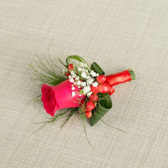 Set against a gently textured, light beige fabric that looks like finely woven linen, a horizontal floral boutonniere stretches from the lower left of the frame towards the upper right, as if poised to be lifted and pinned for a special event in North Watford. At its core is a striking fuchsia-pink rose, fully in bloom yet still tight enough to show a luxurious spiral of petals, each one smooth and velvety, holding a deep, romantic colour that immediately draws the eye. Surrounding this central rose, tiny white flowers reminiscent of baby's breath gather in small clouds, their delicate, star-like blooms lending softness and a hint of bridal tradition, ideal for weddings near Cassiobury Park or around the High Street. Bright red hypericum berries are positioned carefully among the whites and greens, their round, glossy forms adding both festive colour and a playful change in shape, like little jewels nestled in the foliage. Fine, feather-like sprays of green, possibly asparagus fern, extend outwards in graceful arcs, adding lightness and movement, while darker, broader leaves are skilfully curled and woven to form a structured cradle that supports the entire arrangement. The stems at the base are neatly and tightly bound in a lustrous red satin ribbon, the fabric wrapped smooth and finished in a tidy knot that feels secure yet refined. The overall impression is of a meticulously designed boutonniere or small corsage, radiating romance, joy, and polished elegance, perfectly suited to a groom, groomsman, or any formal guest arriving at a celebration and looking for that final, meaningful floral detail.
