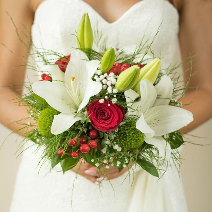 A detailed close-up presents a radiant bride from North Watford, her hands gently clasping a lavish and thoughtfully composed bridal bouquet against the flowing lines of her elegant, patterned white gown. The bouquet radiates timeless romance, with glowing white oriental lilies at its heart-their petals unfurled in full bloom or curled in fresh green buds, exuding an aura of innocence and devotion. Tucked among them is a striking deep red rose, tightly furled and luminous, a traditional emblem of romantic passion. Accents of vivid red hypericum berries lend a festive sparkle, while spherical moss-green clusters-likely green trick dianthus-introduce playful texture and a nod to the lush spaces around Cassiobury Park. Wispy baby's breath floats between the larger flowers, adding airy softness and a dreamlike quality, as if carried in from a morning stroll along the River Colne. The foundation of elegant ferns and lush green leaves provides visual fullness and an organic charm that reflects the natural beauty cherished throughout North Watford. Illuminated by soft, diffused light, the arrangement feels both contemporary and classic, an ideal floral choice for weddings graced with local character and tradition. This bouquet doesn't simply match a dress-it weaves together layers of love, anticipation, and artistry, making it a centrepiece to be treasured long after the vows have been spoken.