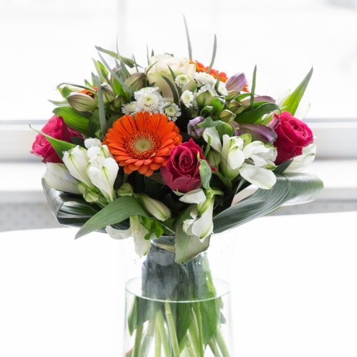 This exquisite North Watford bouquet showcases a harmonious balance of colour and artistry, with each bloom chosen for its lush vibrancy and emotive presence. At the heart sit radiant gerbera daisies: two a vivid, zesty orange, two a gentle, creamy peach, each flower brimming with detail down to its deep brown centre. Rich fuchsia roses, their petals tightly layered, introduce passion and depth that recall the bustling energy found near North Watford's shopping parades. Accents of ivory-white alstroemeria and soft pink buds weave through the composition, offering moments of gentle brightness that evoke the peaceful green spaces of Woodside. The bouquet is cradled by broad, glossy, paddle-shaped leaves-folded with purpose and finesse-circling the stems in a lush, verdant collar that gives the piece a rounded, substantial presence. In between, fine sprigs of foliage and pointed green leaves fill out the arrangement in a way that feels fresh, organic and abundant. Housed in a clear glass vase, the stems' natural green tones shine through, underscoring the freshness and quality promise of a local florist. The bouquet stands illuminated against a soft, light-drenched background, focusing all attention on the intricate blend of colour, texture, and shape. This composition radiates optimism, warmth, and the quiet celebration found in everyday North Watford life-ideal for marking birthdays, anniversaries, or simply extending a thoughtful gesture to someone special.