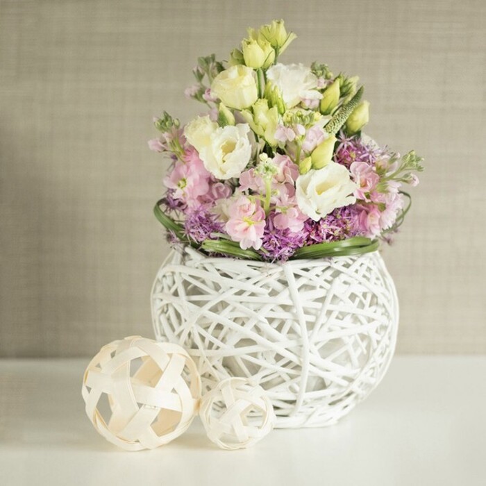In a sunlit corner of North Watford, a beautifully orchestrated floral arrangement is placed atop a pristine white table, offering a visual feast of pastel tones and layered textures. Creamy white and soft yellow eustoma blossoms dominate the bouquet, their gently ruffled petals cascading gracefully among spikes of pale pink stock flowers that lend vertical drama and delicate movement. Beneath these star blooms, a bed of small, vibrant purple filler flowers-possibly aster or early allium-adds a lovely density and contrast, reminiscent of wildflowers glimpsed in local North Watford gardens. Deep green, glossy foliage encircles the base, framing the bouquet and lending it structure. The arrangement is thoughtfully presented in a contemporary, rounded white vase, constructed from an open lattice of painted rattan or dried branches-a nod to North Watford's blend of modern design and traditional craft. Next to the vase, two decorative spheres made from lighter rattan rest on the surface, their open basket-weave pattern echoing the vase's tactile qualities and bringing an extra touch of natural character. All elements sit before a soft beige or greige wall, the subtle fabric-like texture enhancing the overall serenity and giving the flowers centre stage. Capturing the freshness and elegance so often associated with springtime across North Watford, this design perfectly suits wedding tables, anniversary celebrations, or simply brightening a family home, delivered with loving expertise by North Watford's local florists.