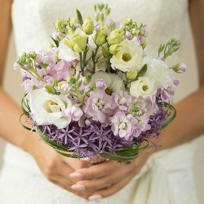 A delicate, close-focus image draws our attention to a sophisticated bridal bouquet being tenderly cradled by the hands of a North Watford bride. Light pours in softly, illuminating the bouquet's lush, rounded form: prominent creamy-white lisianthus blossoms unfurl in gentle, ruffled waves, their subtle yellow-green centers shining with understated brilliance. These blooms are interlaced with slender, unopened lisianthus buds and airy clusters of multi-petaled pink and lilac stock, introducing gentle texture and a sense of movement throughout the arrangement. At the base, a collar of deep violet, star-shaped allium flowers encircles the bouquet, providing dramatic structure and unique contrast-much like those found in the gardens of North Watford or Cassiobury. Artistic dark green bear grass is woven with care above the allium, looping in elegant arcs to frame the design. The bride's hands-nails polished in a whisper of blush-cup the partially visible, ribbon-wrapped stems, adding further grace to the arrangement. In the softly blurred background, the white of a wedding gown, perhaps detailed with lace, further enhances the bouquet's pristine color palette while ensuring all focus remains on the craftsmanship and freshness of the flowers. This wedding bouquet, ideal for celebrations in North Watford or Watford's leafy surroundings, beautifully bridges classic romance and modern floral artistry, making it a prized choice for local florists and brides alike.