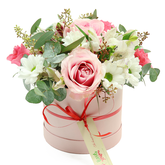 An elegantly styled hatbox flower arrangement is displayed against a bright white background, its pastel colours and soft textures giving the scene a calm, uplifting feel that suits a thoughtful delivery anywhere in North Watford. The round, cylinder-shaped box is finished in matte blush pink, suggesting a high-quality, contemporary presentation. Wrapped neatly around its middle is a slim satin ribbon in a vibrant coral-red shade, gathered at the front into a tidy bow. A rectangular cream gift tag with graceful green lettering and a small printed website hangs from the bow, subtly indicating a curated floral gift rather than a casual bunch. Above the rim of the box rises a lush, domed bouquet composed of pink, white, and green tones, carefully arranged to look both full and airy. The eye is quickly drawn to one particularly striking rose near the centre: its outer petals are a gentle pale pink, while the inner spiral deepens to a richer hue, creating a romantic, heart-like swirl. Surrounding it, white chrysanthemums with crisp petals and bright yellow-green centres form clean, circular shapes that echo the roundness of the box itself. Slender-stemmed white alstroemeria, recognisable by their elongated petals and fine internal speckles, weave between the larger flowers, softening the structure. Pockets of deeper, fuchsia-pink blossoms-likely small roses or lisianthus-peek through, adding warmth and visual rhythm akin to the colourful doorways and gardens scattered around North Watford side streets. Silver-green eucalyptus leaves with their rounded, coin-like shapes arc gently throughout the bouquet, adding both softness and a contemporary touch, while darker leafy stems and tiny green buds give depth and a slightly wild, garden-grown character. The overall impression is of a lovingly hand-crafted hatbox arrangement, ideal for celebrating birthdays, anniversaries, or Mother's Day, and perfectly suited to be delivered as a charming surprise to homes and offices across North Watford.