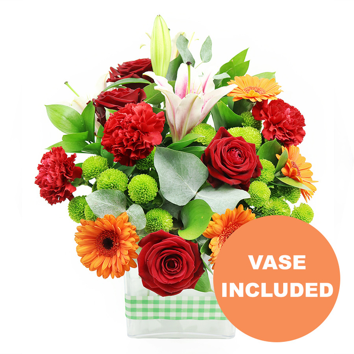 In this detailed product image, a joyful, densely packed floral arrangement is presented in a clear, rectangular glass vase, photographed on a pristine white background that makes every colour sing. Around the lower half of the vase runs a band of light green and white gingham ribbon, bringing a touch of rustic charm that would feel right at home on a dining table in North Watford or in a cosy kitchen overlooking the nearby parks. The bouquet itself is a harmonious blend of warm and fresh tones. Several deep crimson red roses, their petals velvety and spiralling inward, stand out as luxurious focal points. They are flanked by bright orange gerbera daisies, their flat faces and dark central eyes turned forward, creating a burst of sunshine against the cooler greens. Threaded through the roses and gerberas are rich red-orange carnations, each head densely frilled, adding a sense of softness and generous volume, like layers of fine fabric. Towering above and between these blooms are elegant white oriental lilies; some flowers are fully open, with elongated petals marked by subtle pink stripes and prominent stamens, while other buds remain closed, tall and smooth, hinting at blossoms still to come. Pockets of vivid lime-green pompom chrysanthemums nestle between the larger blooms, their rounded, button-like forms providing playful texture and a refreshing, dewy quality. The arrangement is anchored by deep green foliage and sprigs of silver-toned eucalyptus, whose slender leaves bring a cool, airy contrast and gentle fragrance. In the lower right portion of the image, a circular orange label partially overlaps the bouquet, clearly marked with the words VASE INCLUDED in white capital letters, reinforcing that this striking mixed-colour arrangement arrives in its own stylish vase, ready for same-day delivery to addresses throughout North Watford.