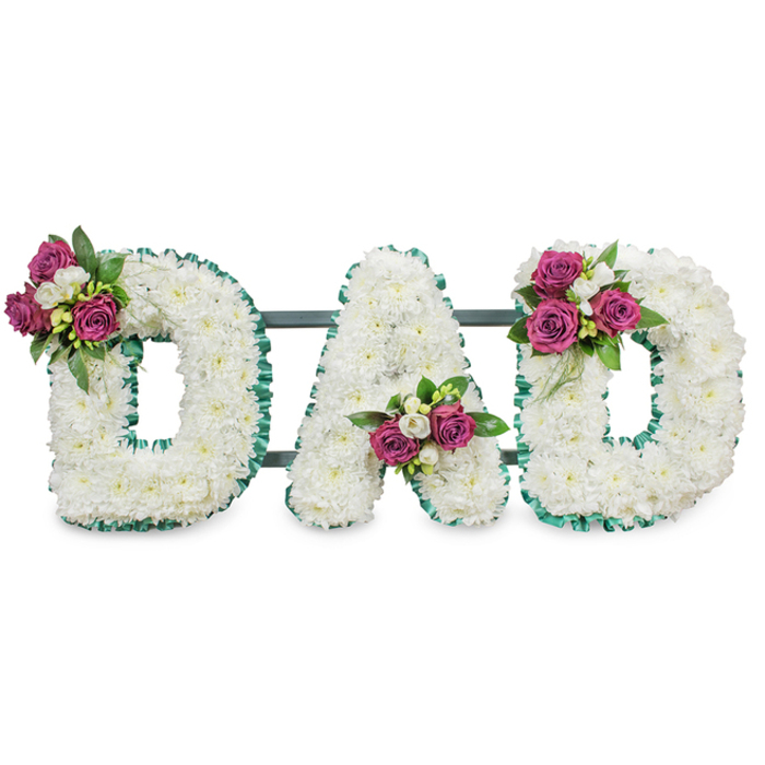 This striking sympathy arrangement spells DAD in large, sculpted capitals, each letter composed of a dense mass of pristine white chrysanthemums that together form a soft, voluminous canvas. The edges of every character are crisply outlined with an emerald green pleated ribbon, its ruffled profile offering a stately border that sets off the immaculate whites. A low, discreet dark green or grey bar supports the composition from behind, indicating a self-standing memorial piece designed for display at funeral services or a graveside. Accent clusters are intentionally placed to create visual balance: the first D bears a bouquet on its upper left curve, the A receives a compact spray tucked into its central lower V, and the second D is mirrored with a cluster on the upper right. Each cluster includes deep magenta roses in mixed stages of bloom, delicate white freesia or similar small white blossoms, and lush dark foliage that gives the arrangement an evergreen depth and a subtle, comforting scent. The floral texture is tactile and the colour palette restrained-white purity, vivid magenta punctuation, and a restful emerald frame-conveying reverence without excess. As a local floral designer working around North Watford and Cassiobury Park, I would present this piece with soft morning light and quiet hands, mindful of the service route between Watford High Street chapels and Vicarage Road venues. It reads as a tender, bespoke expression of grief and honour, created to hold memory and offer solace.