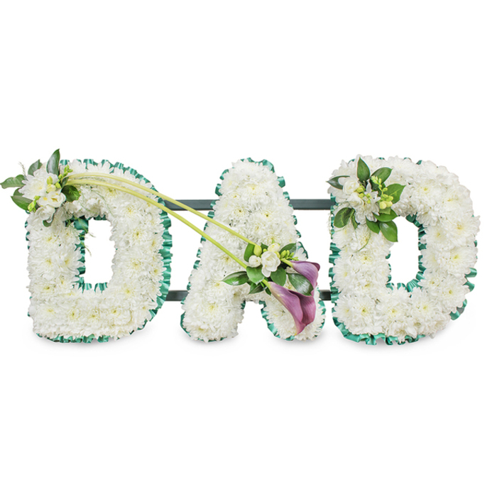 A poignant funeral floral tribute forms the word DAD in large upright letters, a classic memorial design often chosen by families in North Watford to honour a father's memory. Each letter is built on a sturdy frame and densely covered with pristine white chrysanthemums or similar small white blooms, placed so closely together that no gaps in the base are visible. This tight, cushion-like texture creates a serene field of white, softly catching the light against the neutral background. Encircling the outer contour of each letter is a neat band of emerald green ribbon, gently gathered into pleats so the border looks ruffled and slightly raised. This vibrant green edge recalls the fresh lawns and hedgerows around North Watford and provides a vivid contrast that makes the lettering instantly readable, even from a distance in a chapel or crematorium. On the gentle curve of the first D sits a small spray of white freesia and mixed foliage, their delicate buds and open blooms adding a hint of fragrance and a looser, garden-like feel. The final D carries a similar, slightly smaller cluster on its upper right corner, balancing the design. The central A becomes the focal point, where two long green stems sweep gracefully from the first D across the arrangement, leading the eye towards a rich accent of colour: two deep purple calla lilies, cradled among additional white blossoms and greenery. Their velvety trumpets introduce a sense of depth, dignity, and quiet drama. This DAD tribute, ideal for display at a funeral, wake, or graveside service in North Watford, expresses enduring love, thanks, and remembrance through its calm palette and thoughtful detailing.