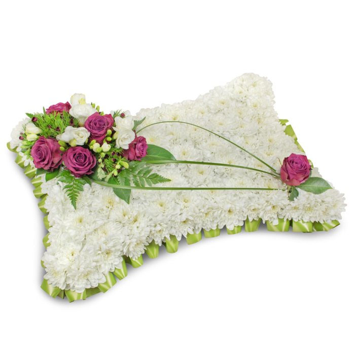 Against a stark white setting this traditional pillow-style sympathy arrangement conveys quiet dignity and comfort. The entire rectangular cushion is densely planted with pristine white chrysanthemum florets; up close, the surface reads like a soft, quilted field of petals that absorbs light and suggests a tranquil stillness. A prominent cluster sits in the top-left corner: several deep fuchsia or magenta roses, their velvet petals unfurling in measured layers, are woven with small white freesia blooms that add a whisper of perfume and fragile contrast. Surrounding greens-fronded fern and assorted glossy leaves-frame the accent cluster and anchor the colour, while three long, dark-green bear grass blades sweep in elegant arcs across the chrysanthemum bed, introducing a deliberate, calming line. Toward the bottom-right a solitary magenta rose mirrors the main cluster, tucked among leaves to achieve a balanced, contemplative composition. The pillow's edge is finished in a folded light-green satin ribbon, pleated into a neat scallop that gives the tribute a refined border and a hint of warmth against the cool white. The overall impression is one of rest and respectful remembrance: soft to the eye, faintly perfumed, and carefully proportioned for presentation at funeral services or memorials in North Watford-often chosen by families arranging tributes near Watford High Street or local funeral homes seeking a classic expression of sympathy.